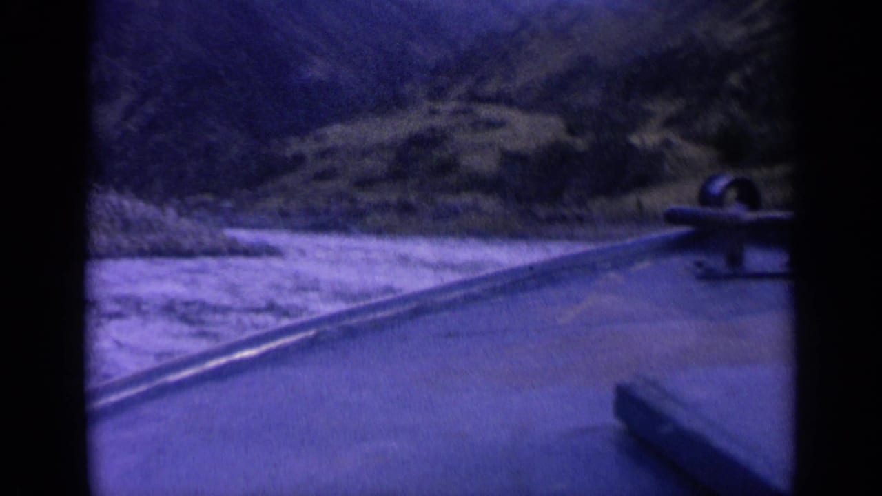 Blue-purple 1963 Washington 8mm frame showing wake across the water, a small boat ahead, and rugged slopes around the channel.