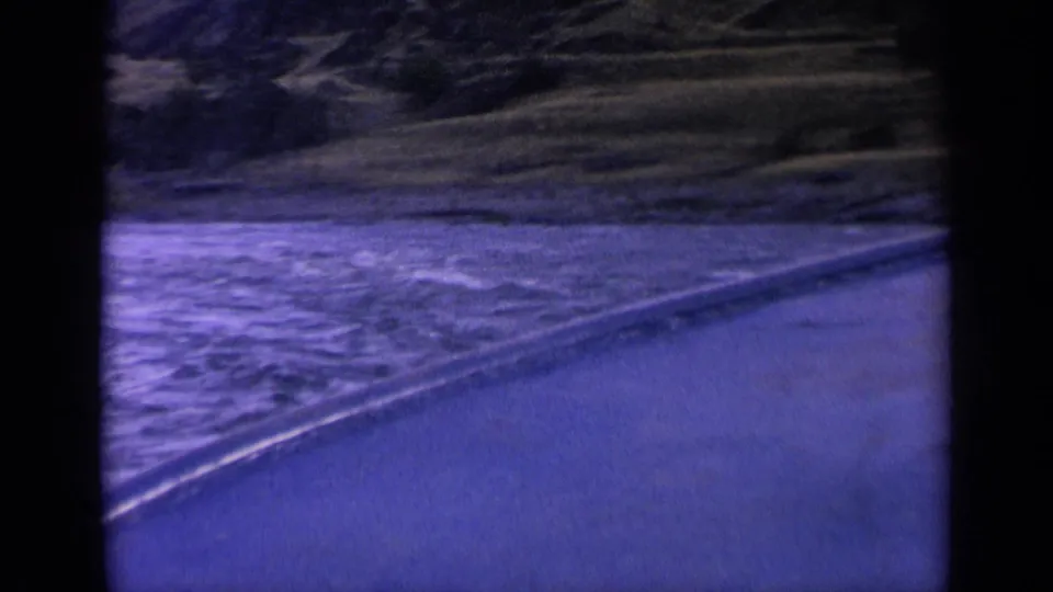Blue-shifted 8mm view of water passing a curved embankment with wooded hillside terrain in the background.