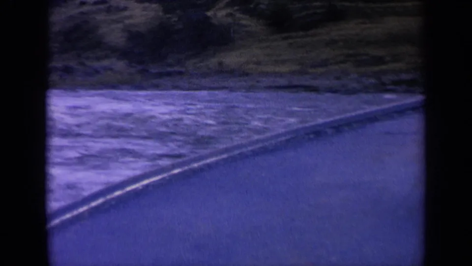 Soft 8mm frame of a Washington waterside landscape, showing blue-tinted water, a curved bank, and wooded hills.