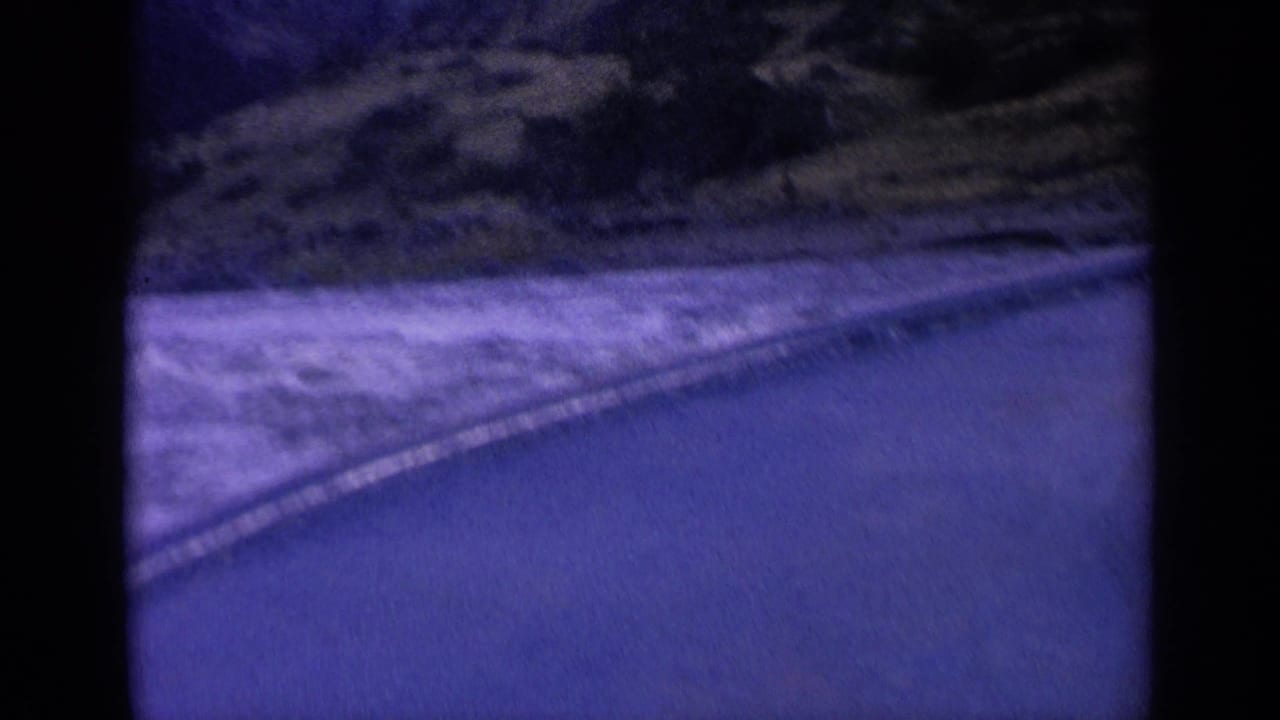 1963 Washington 8mm frame showing turbulent water beside a roadside, viewed from a moving vehicle with a purple-blue cast.