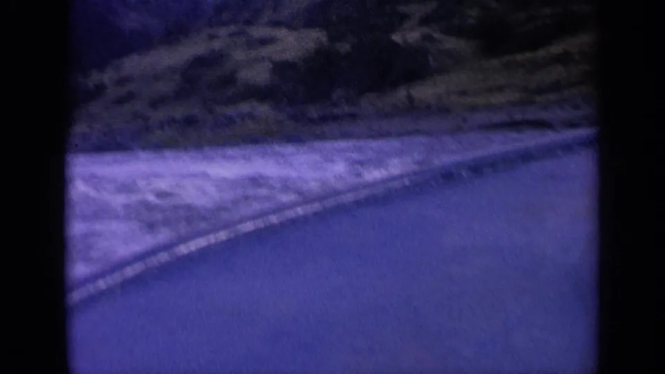 1963 Washington 8mm frame showing turbulent water beside a roadside, viewed from a moving vehicle with a purple-blue cast.