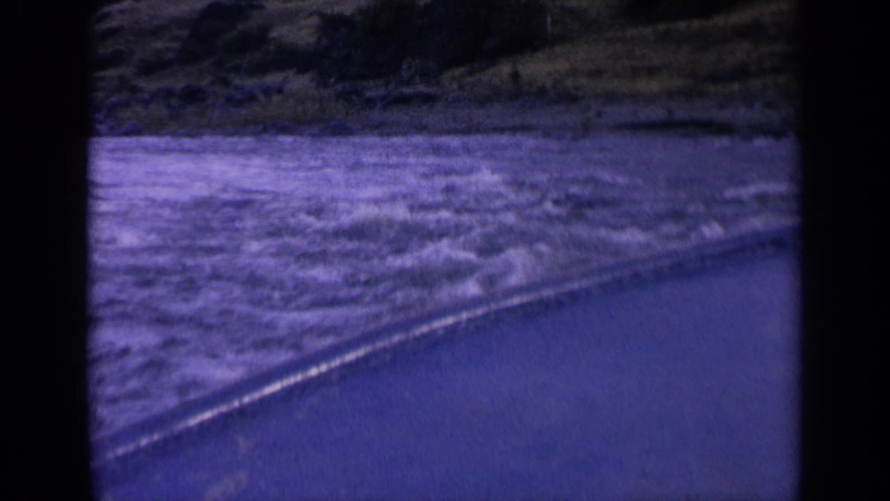 Blue-shifted 1963 Washington 8mm frame showing foamy water near a road, with the camera moving parallel to the shore.