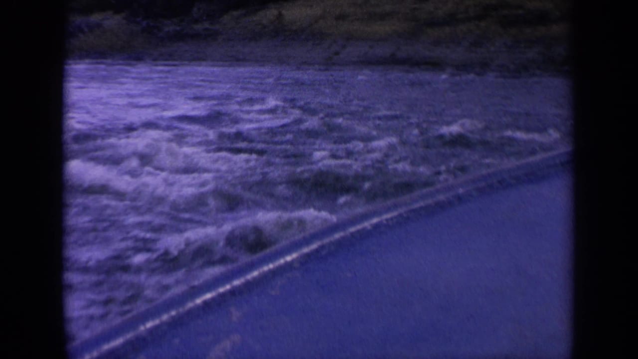 1963 Washington 8mm frame with rough water, diagonal roadside edge, and hillside background seen from a moving vehicle.