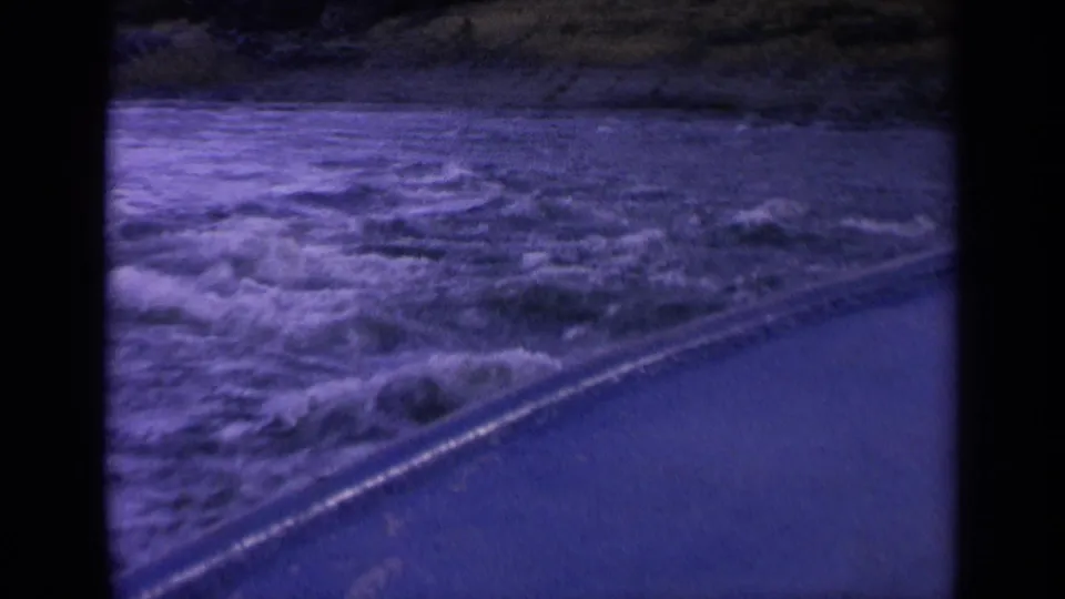 1963 Washington 8mm frame with rough water, diagonal roadside edge, and hillside background seen from a moving vehicle.