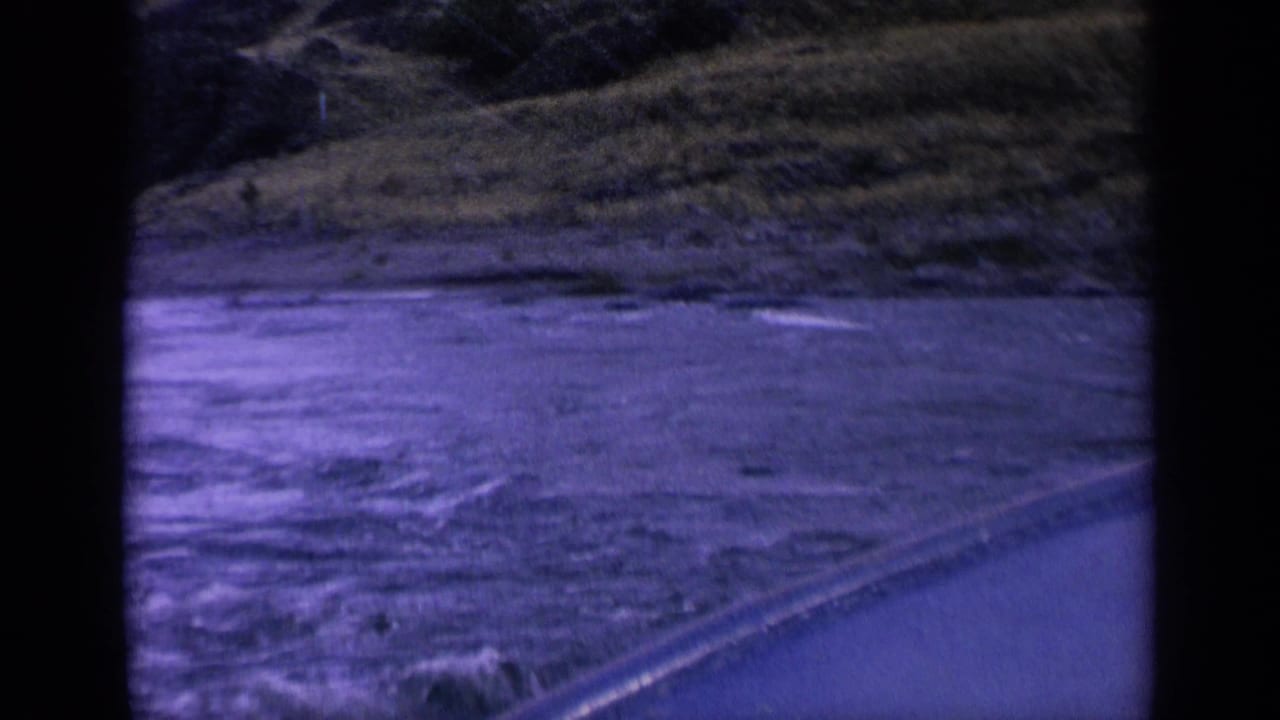 1963 Washington 8mm frame showing foamy moving water, shoreline vegetation, and the diagonal edge of a boat or embankment.