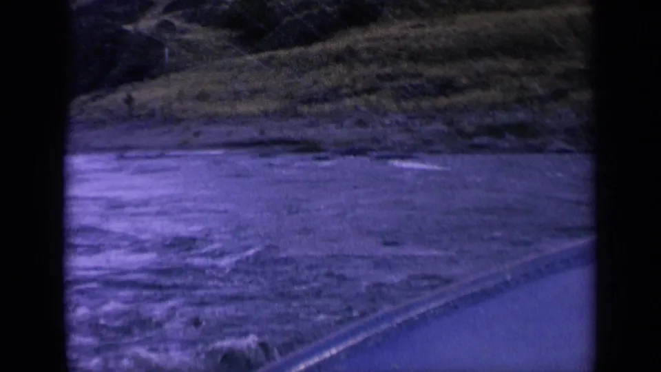 1963 Washington 8mm frame showing foamy moving water, shoreline vegetation, and the diagonal edge of a boat or embankment.