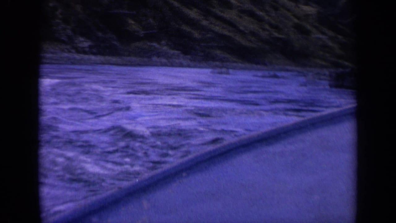 1963 Washington 8mm frame showing churning blue water, a sloped vessel edge, and a dark rocky bank in the background.