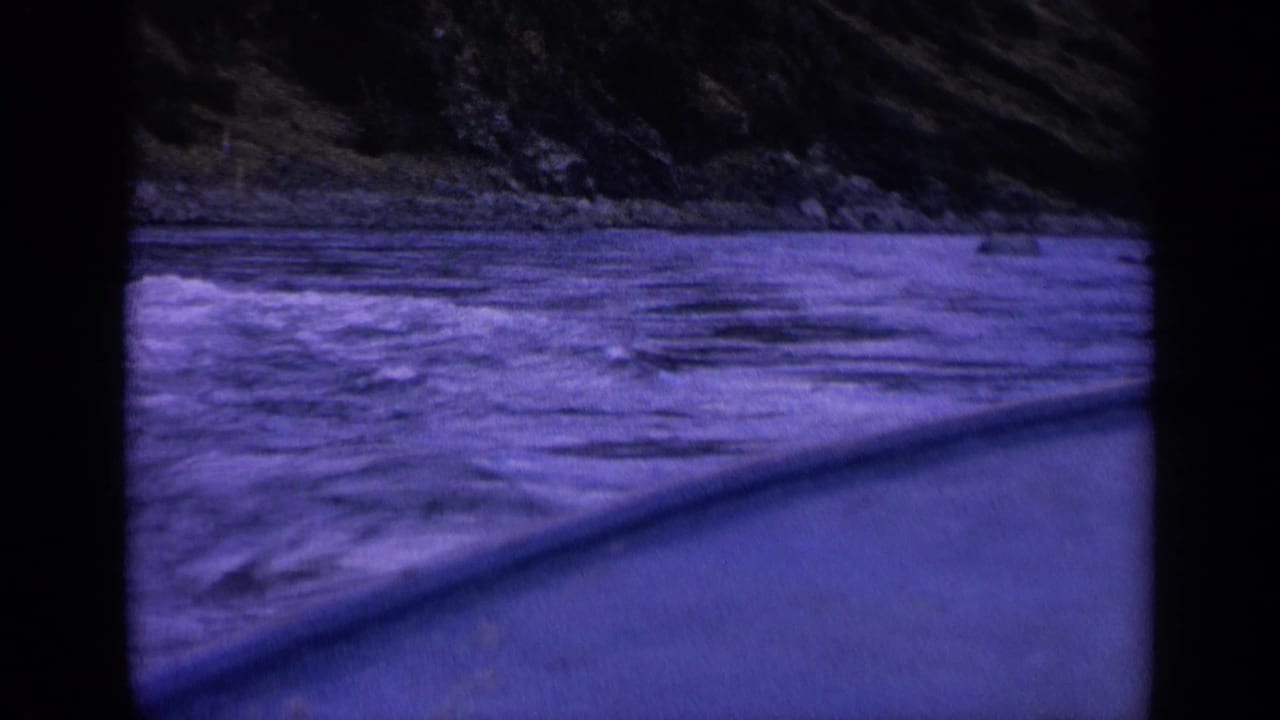 Blue-purple 8mm view of rough Washington water beside a slanted vessel edge, with a rocky bank across the channel.