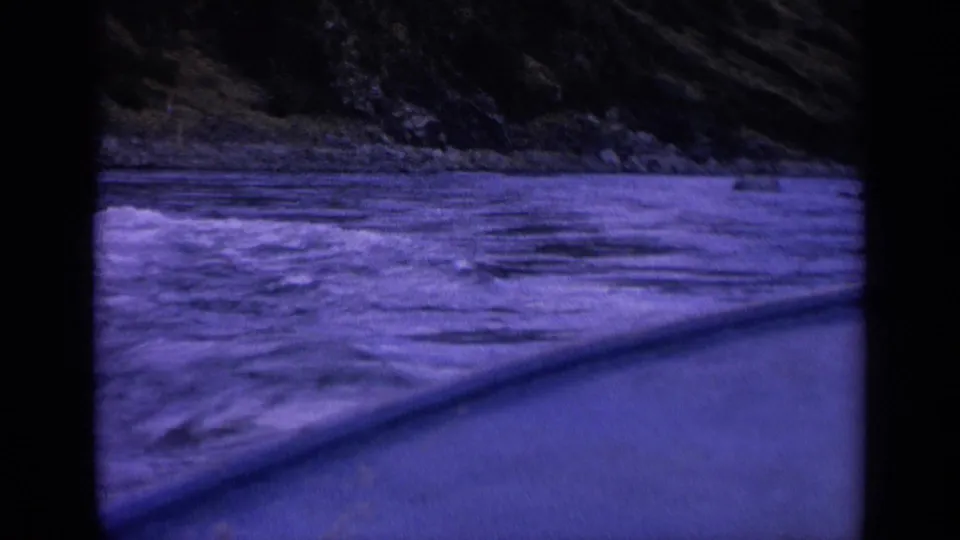 Blue-purple 8mm view of rough Washington water beside a slanted vessel edge, with a rocky bank across the channel.