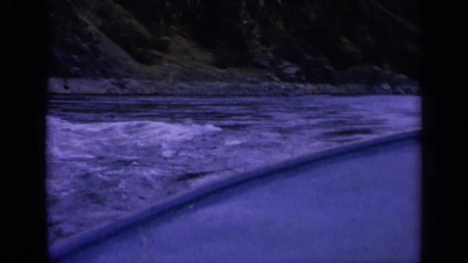 Grainy 1963 Washington home-movie frame with blue water, a dark rocky shoreline, and a sloped boat or window edge.
