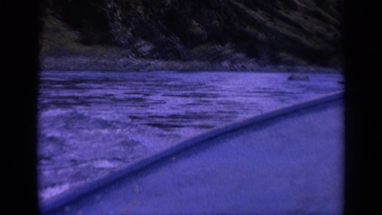 1963 Washington 8mm home-movie frame from a small boat, looking across rippling river water toward rocks and a steep wooded bank with dark film borders.