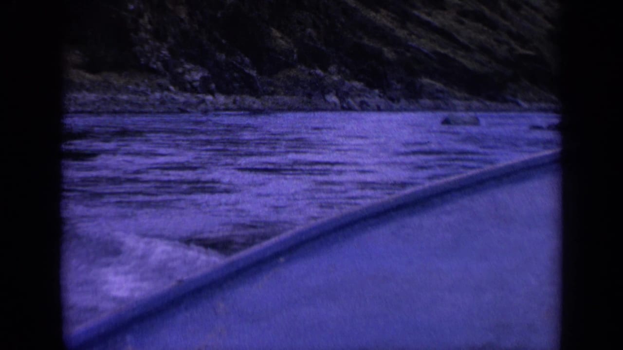 Boat-edge perspective on a Washington river in 1963, with dark rippling water, low rocks along the shoreline, and a wooded slope beyond the 8mm frame border.