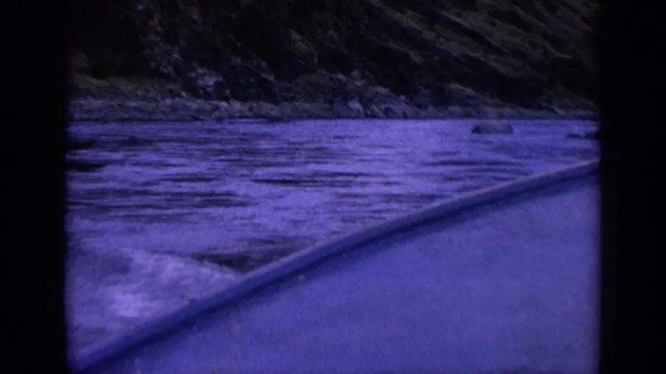 Boat-edge perspective on a Washington river in 1963, with dark rippling water, low rocks along the shoreline, and a wooded slope beyond the 8mm frame border.