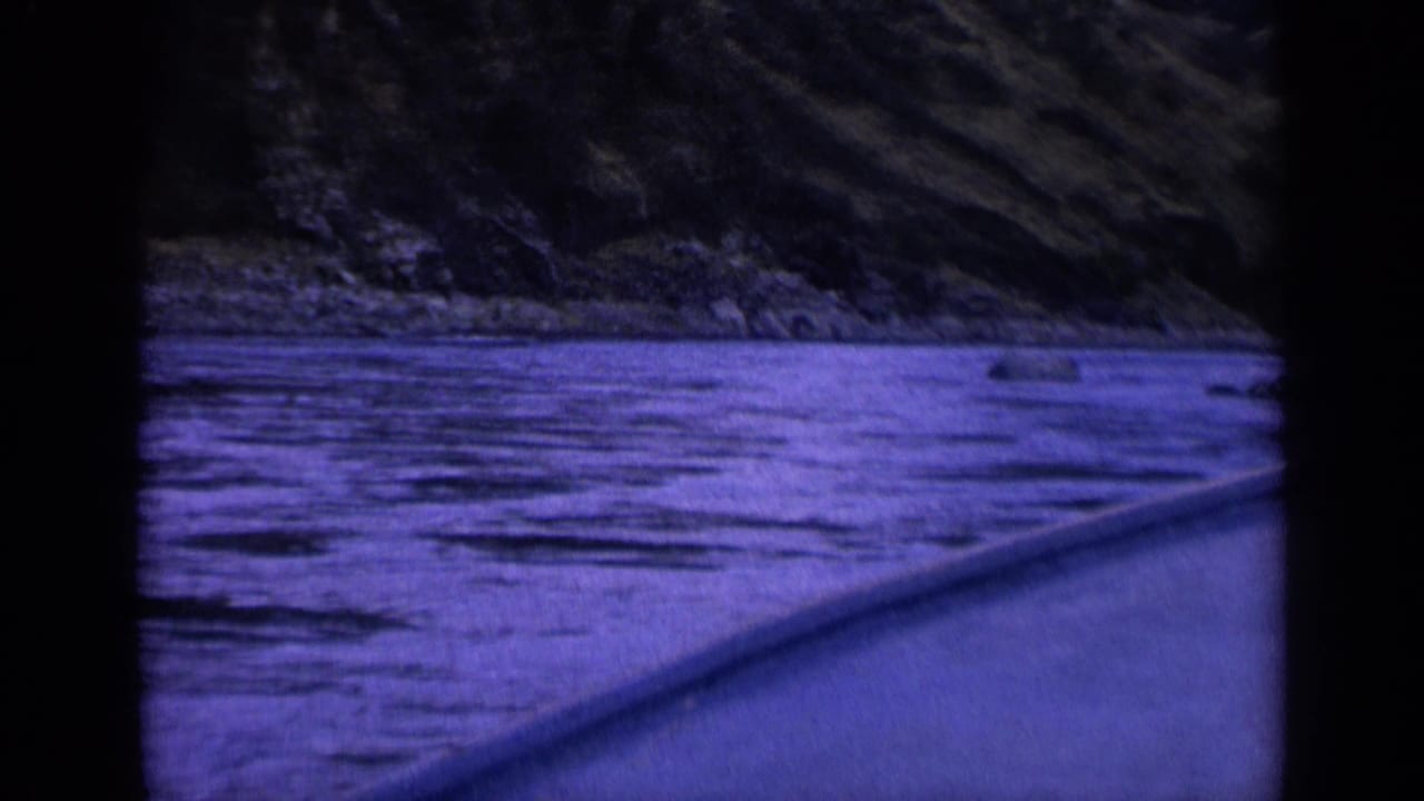 A handheld 8mm frame shows the boat gliding beside a rippling Washington river, with a rocky shoreline and dark wooded hillside in the background.