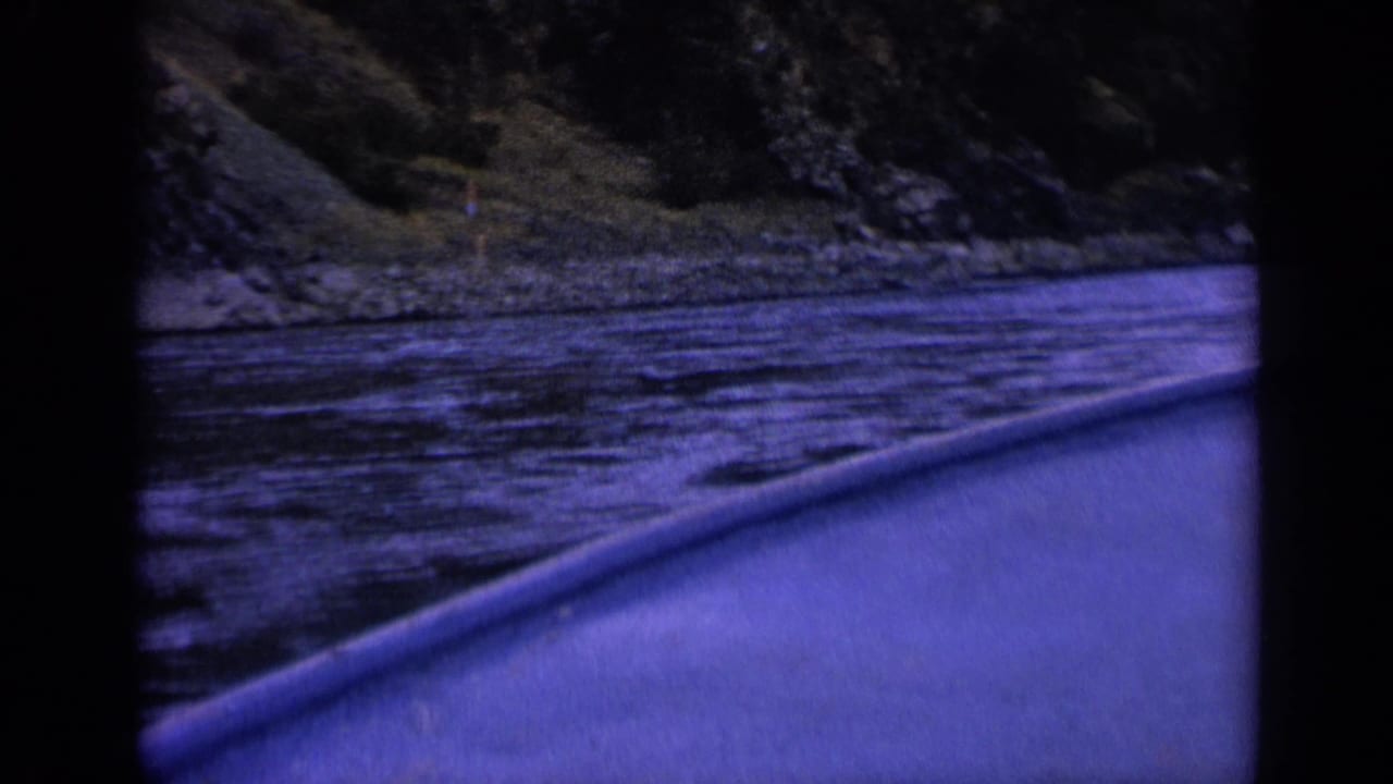 Final 1963 Washington river frame from a small boat, showing the boat side, rippled water, shoreline rocks, and a wooded slope under a pronounced purple-blue film cast.