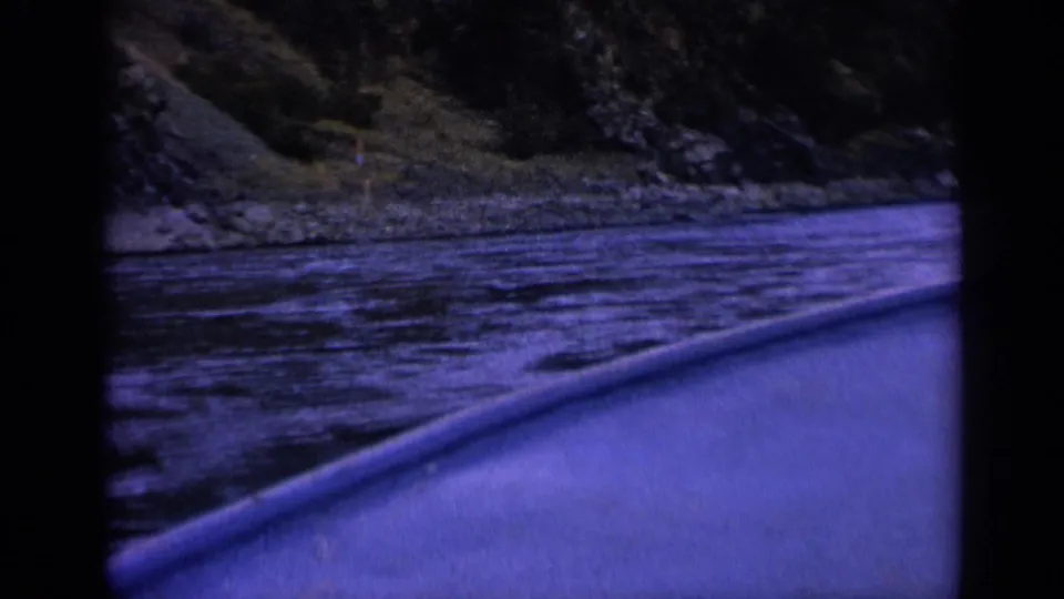 Final 1963 Washington river frame from a small boat, showing the boat side, rippled water, shoreline rocks, and a wooded slope under a pronounced purple-blue film cast.