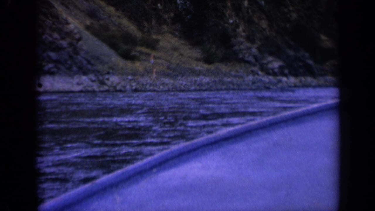 1963 Washington 8mm boat POV looking across rippled river water to a rocky, tree-lined bank with a blue-purple film cast.
