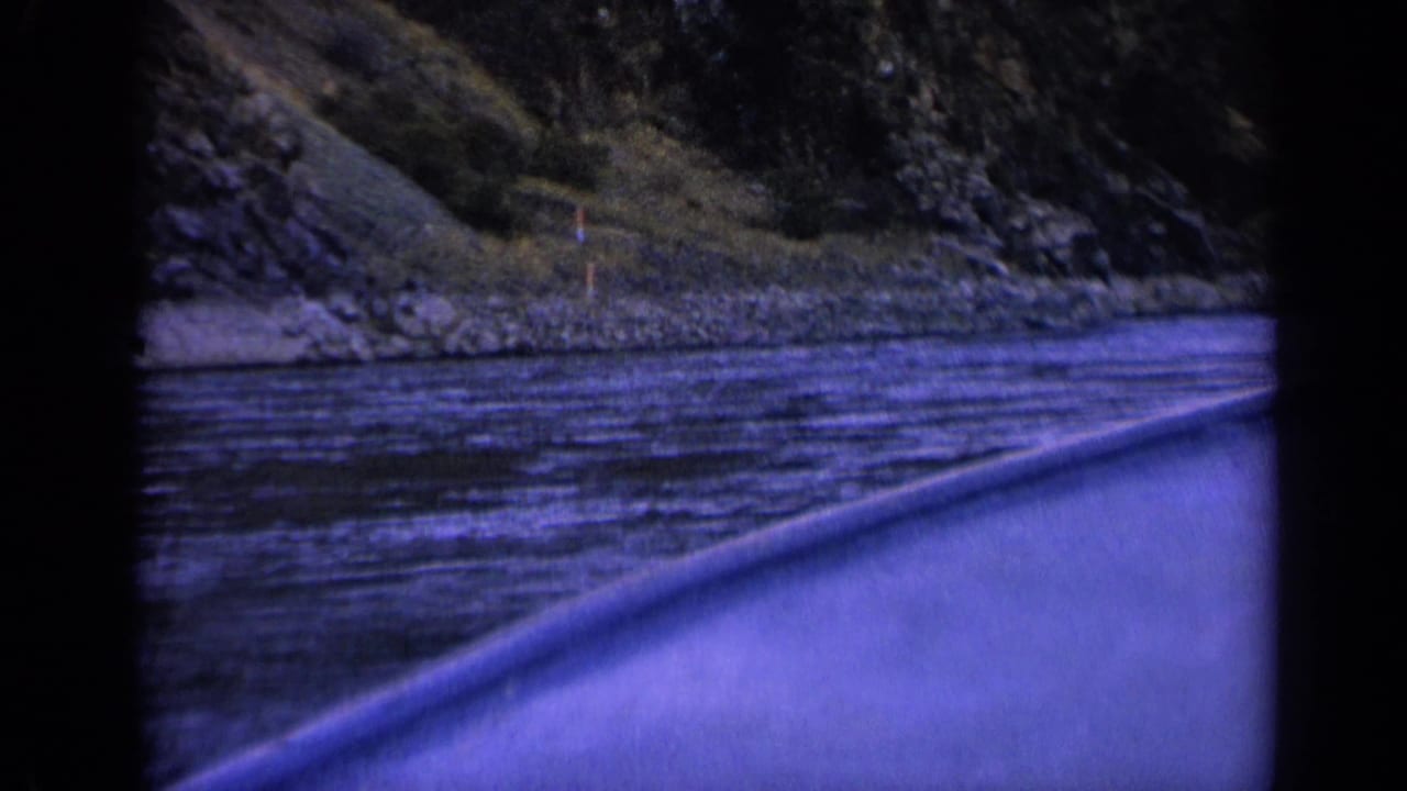 1963 Washington 8mm frame from a moving boat, with the bow or rail at right and a wooded rocky riverbank ahead.