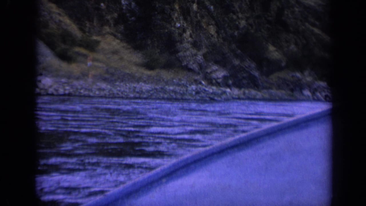 Blue-purple 8mm boat POV on a Washington river, showing rippled water, a rocky bank, and dense trees in soft focus.
