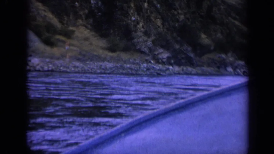 Blue-purple 8mm boat POV on a Washington river, showing rippled water, a rocky bank, and dense trees in soft focus.