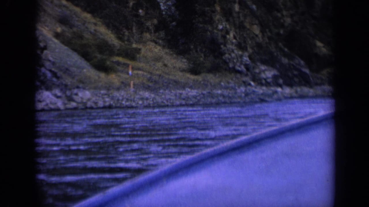 1963 Washington home-movie frame from a boat, showing a riverbank with rocks, trees, and a small upright marker or post near shore.