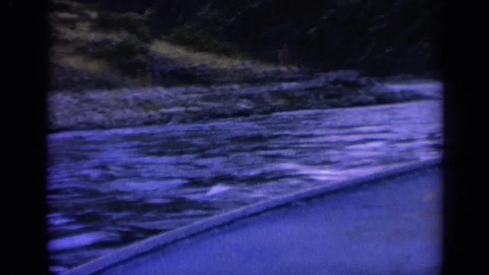 1963 home-movie frame of a riverbank and mountain slope viewed from a moving vehicle in Washington.