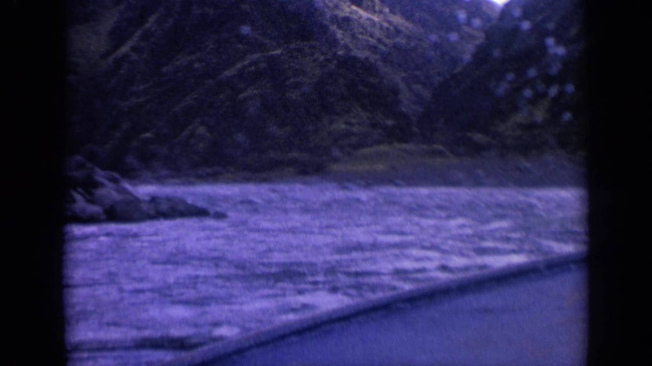 Vintage 8mm frame of a mountain waterway from a moving boat, with rocky shore visible at left.