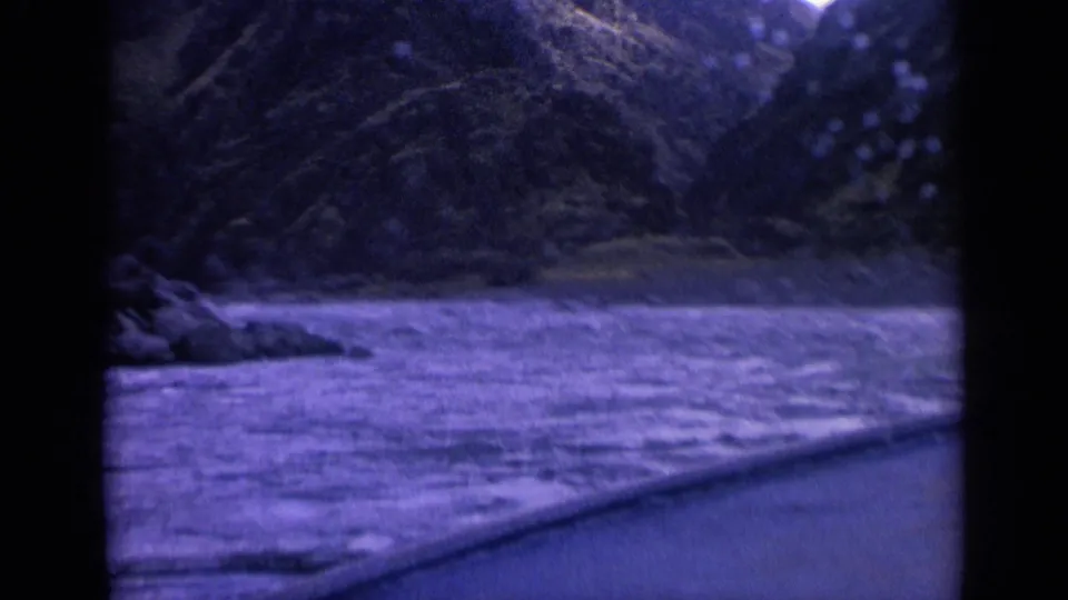 Vintage 8mm frame of a mountain waterway from a moving boat, with rocky shore visible at left.