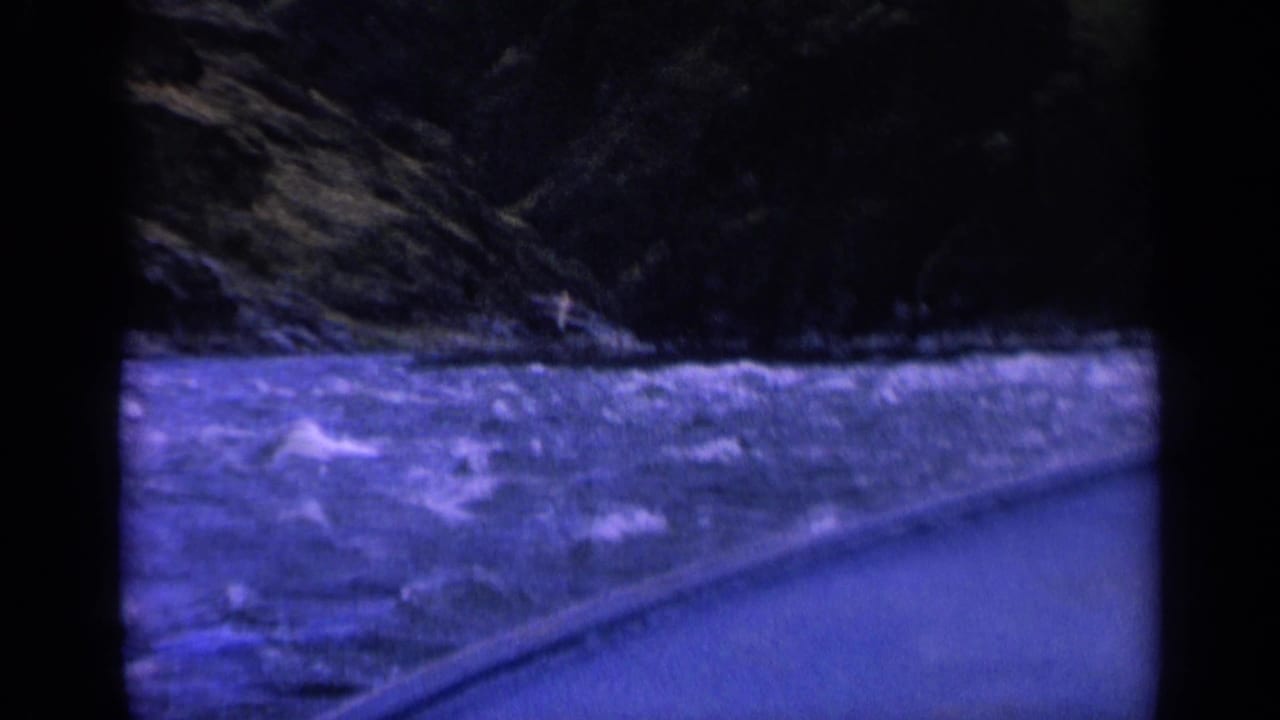 1963 Washington 8mm frame showing rough blue river water beside a paved roadside under dark forested hills.