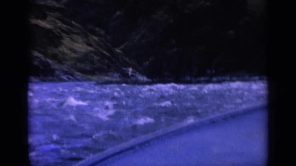 1963 Washington 8mm frame showing rough blue river water beside a paved roadside under dark forested hills.
