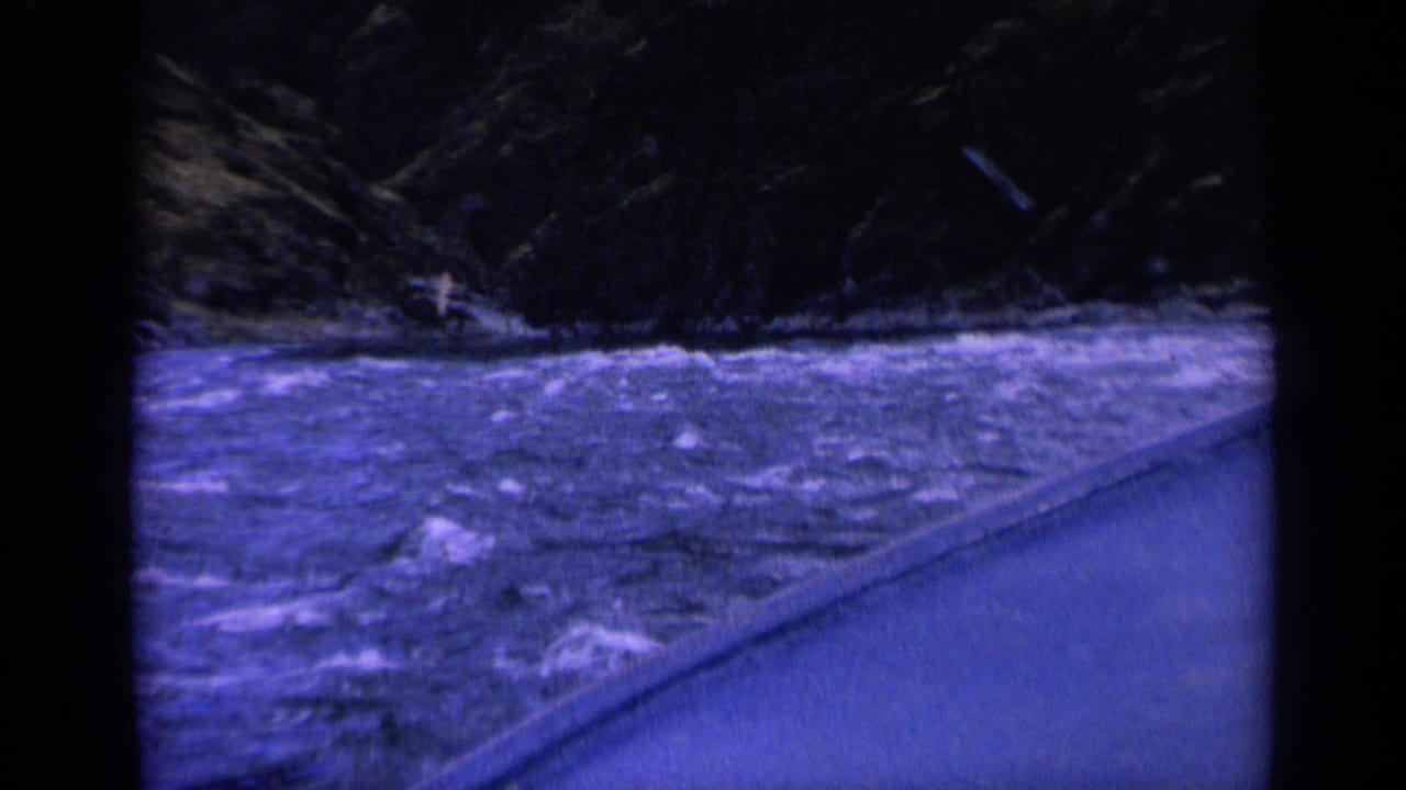 Rough river waves beside a road with a small distant craft or figure near the far bank.