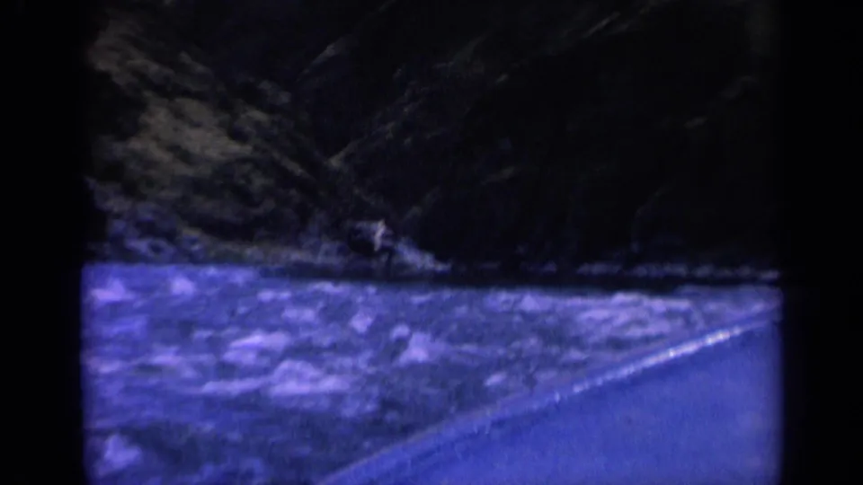 1963 Washington 8mm frame of turbulent water beside a road, with a tiny distant figure or craft near the shadowed far bank.