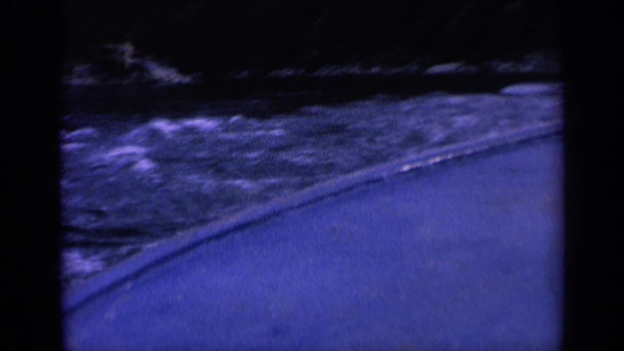 Blue-tinted view over rough river water beside a paved edge and dark shoreline.