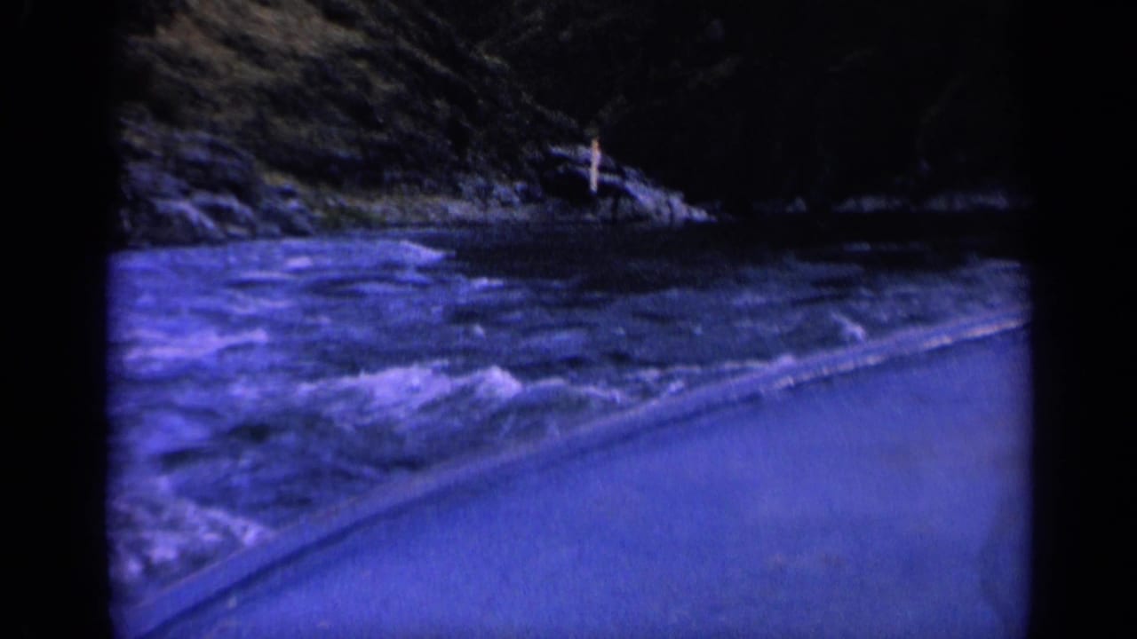 Rough water curves along a paved edge with rocky wooded shore in the background.