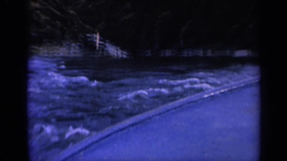 1963 8mm Washington view from a moving boat shows foaming rough water beside a paved bank and wooded shoreline.