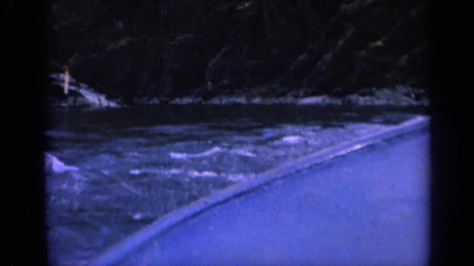 Blue-tinted 1963 Washington home-movie frame showing choppy river water, a paved edge, wooded hillside, and rocky shore.