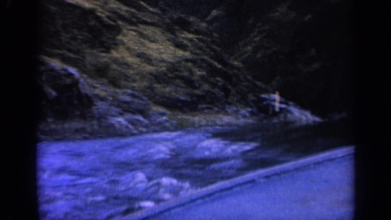 1963 home-movie frame of a boat traveling along a Washington river, with small rapids, hillside, and shoreline marker visible ahead.