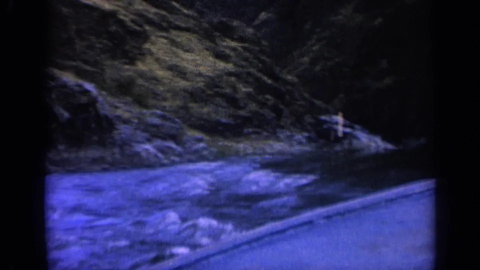 1963 home-movie frame of a boat traveling along a Washington river, with small rapids, hillside, and shoreline marker visible ahead.