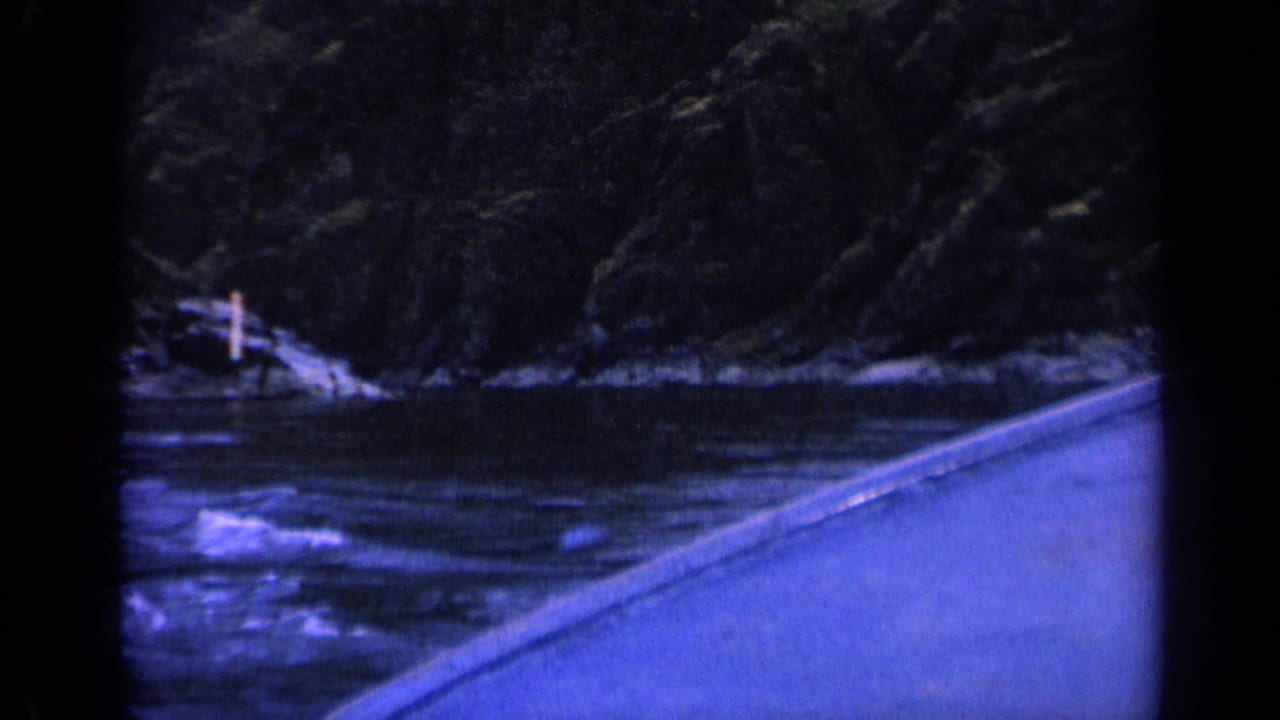 8mm Washington river view from a moving boat, showing dark rocky slopes, flowing water, and a small white marker on the bank.