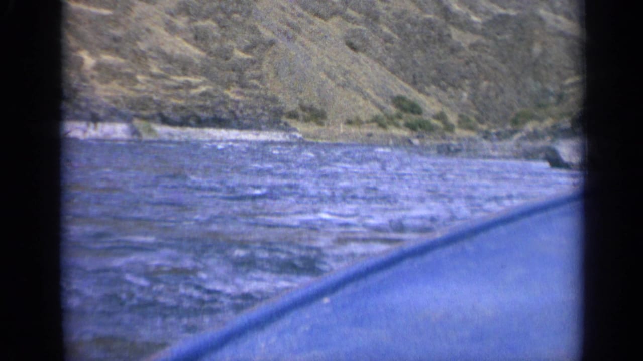 1963 Washington 8mm frame showing blue river current, steep rocky shoreline, and the edge of a boat or canoe in the foreground.