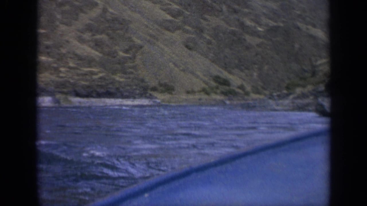 1963 Washington 8mm frame showing a low boat-level view across river current toward a rocky bank and muted mountain slope.