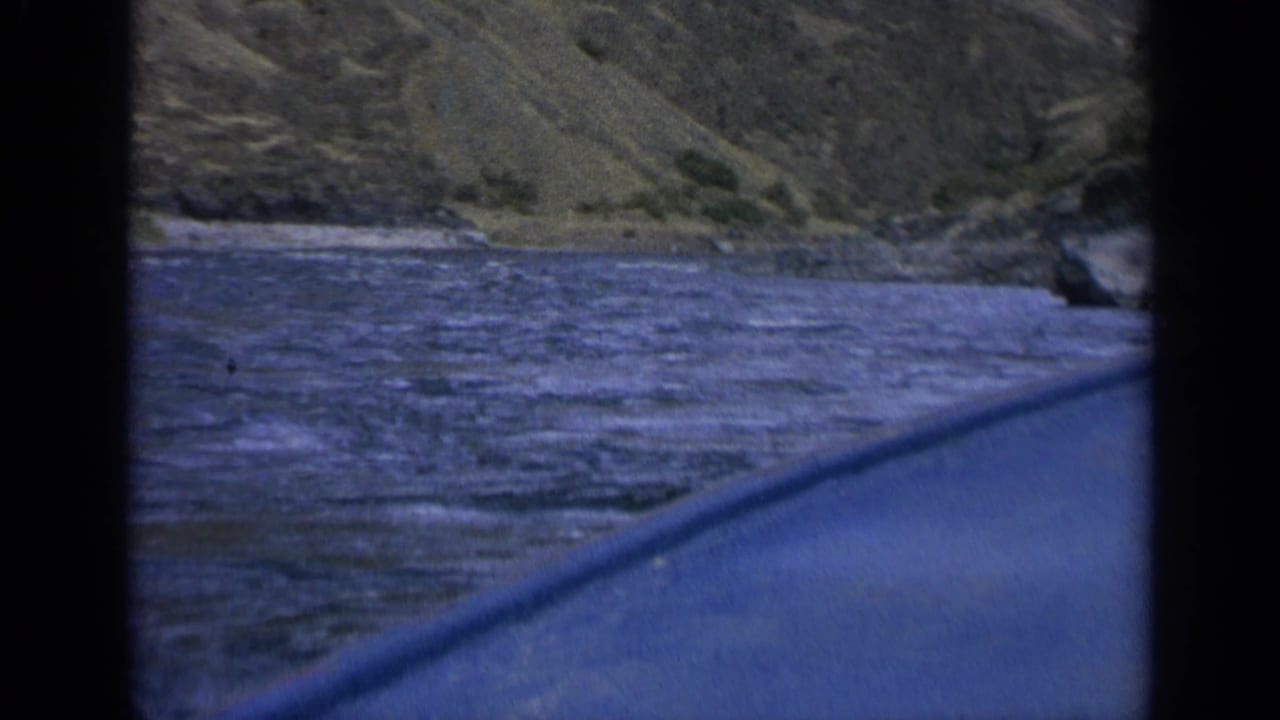 1963 Washington home-movie frame of river current beside a rugged bank, filmed from a boat or canoe with dark frame edges visible.