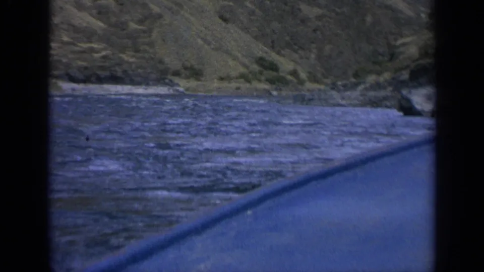 1963 Washington home-movie frame of river current beside a rugged bank, filmed from a boat or canoe with dark frame edges visible.