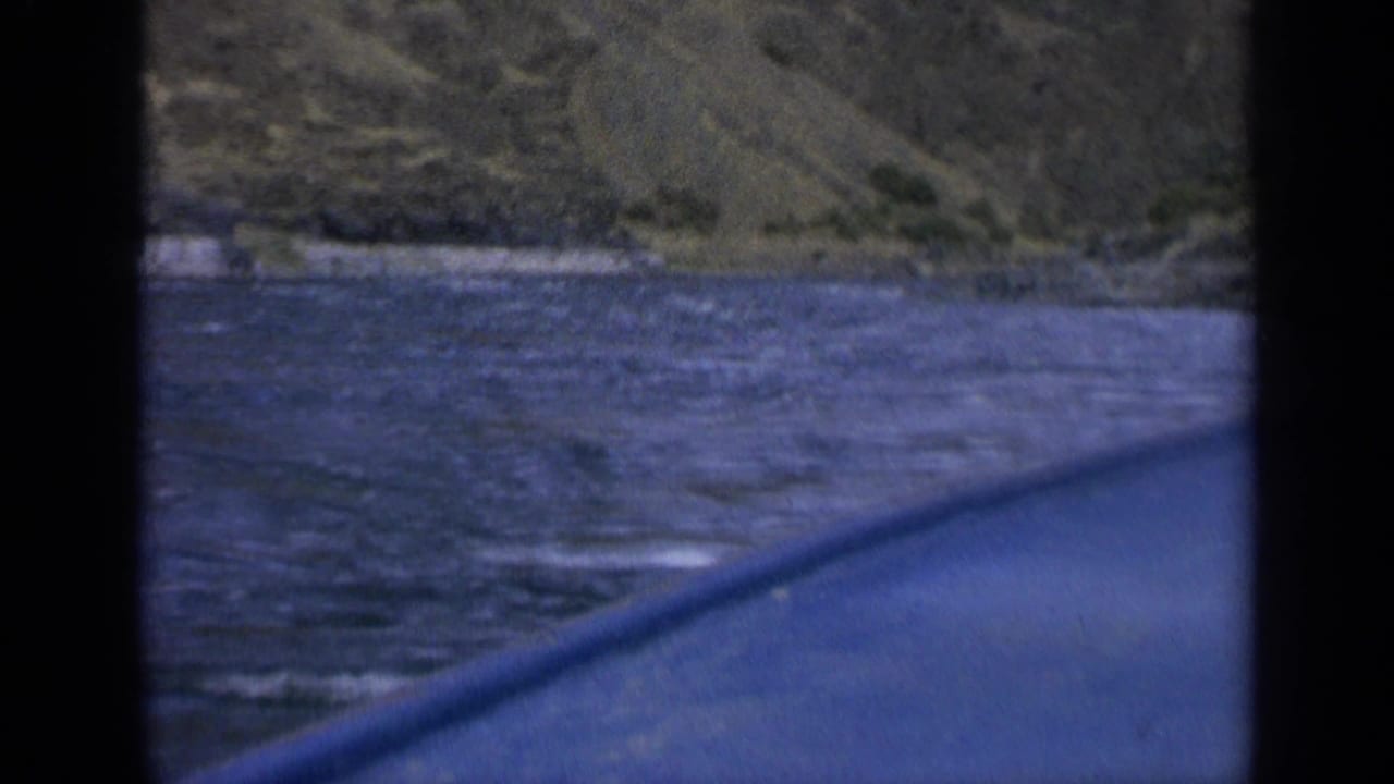 1963 Washington 8mm frame showing rippled river water, a rocky mountain slope, and a blue vessel edge through dark framing.