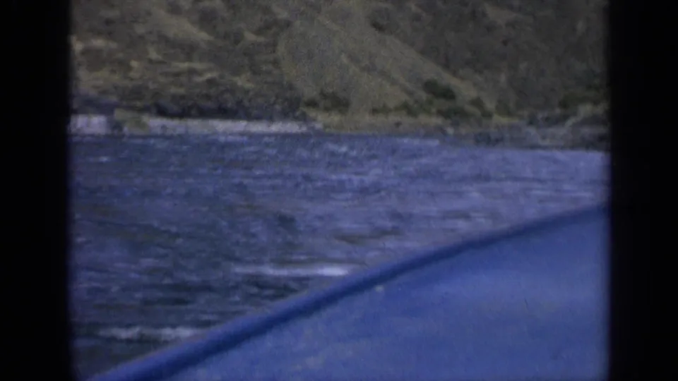 1963 Washington 8mm frame showing rippled river water, a rocky mountain slope, and a blue vessel edge through dark framing.