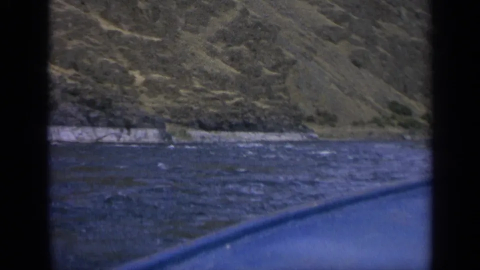 1963 Washington travel footage frame with rippled water, a pale rocky bank, and dry hillside beyond a dark window-like border.