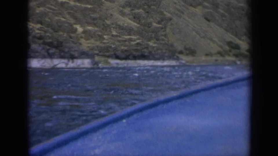 1963 Washington 8mm frame showing river water, shoreline rock, hillside terrain, and the curved blue edge of a vessel.