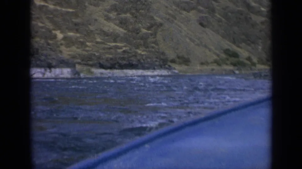 1963 Washington home-movie frame of a rippled river, rocky bank, steep hillside, and blue boat edge through dark framing.