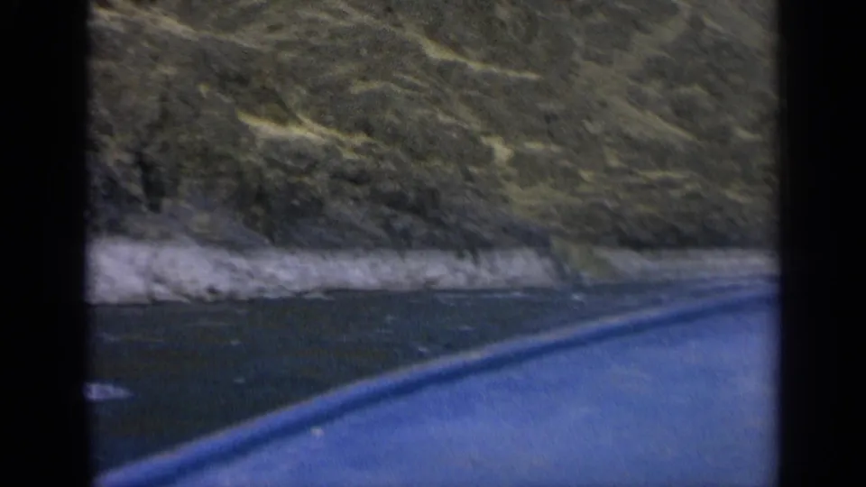 1963 Washington home-movie frame with the blue boat in the foreground and a rugged riverside mountain slope across the water.