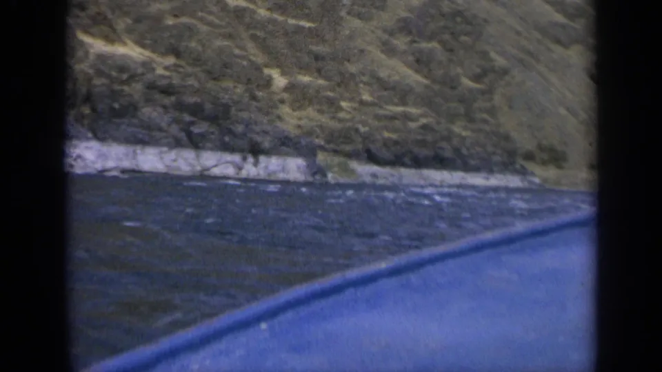 1963 Washington 8mm frame showing a moving boat viewpoint across dark water toward a pale rock shoreline and steep hillside.